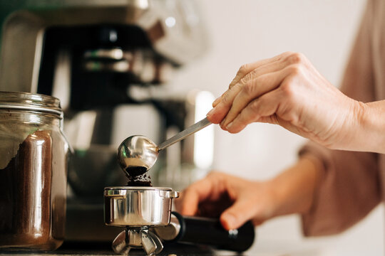 Unrecognizable female with spoon pouring ground coffee into portafilter while standing at kitchen counter with jar of coffee and coffee machine on blurred background