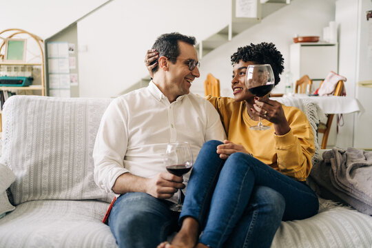 Content multiracial couple chilling on sofa at home with red wine in glasses while enjoying weekend at home and looking at each other