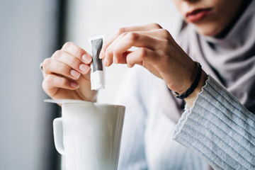 Crop anonymous Muslim female in hijab pouring sugar from paper packet in cup with hot drink in cafe