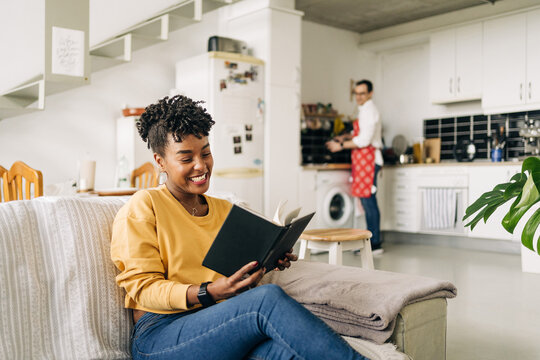 Delighted African American female reading interesting book on sofa on background of man cooking in kitchen