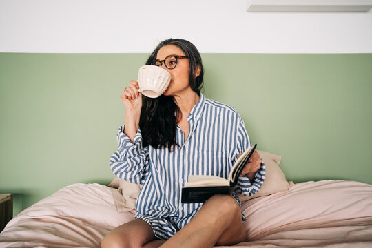 Clever female in eyeglasses with long hair and notebook looking away and resting on soft bed in bedroom