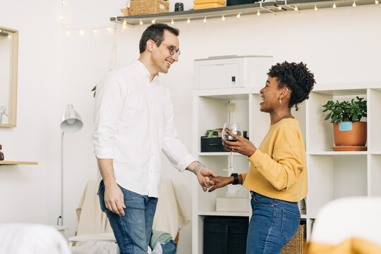 Side view of smiling multiethnic couple in love standing with glass of wine and holding hands while spending romantic weekend at home and looking at each other - Powered by Adobe