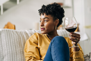 Content African American female sitting on sofa with glass of red wine and enjoying weekend at home while looking away