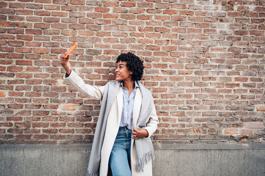 Positive African American female in casual clothes and coat smiling happily while taking self portrait on mobile phone