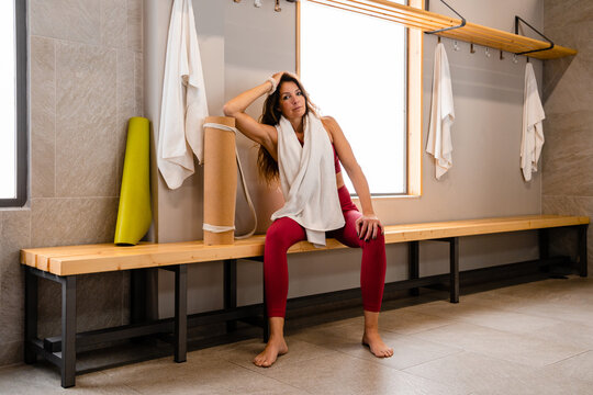 Tired sportswoman in sportswear sitting with towel on shoulders on wooden bench in gym locker room after training