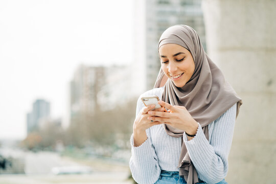 Young ethnic female in hijab standing in city and messaging on mobile phone