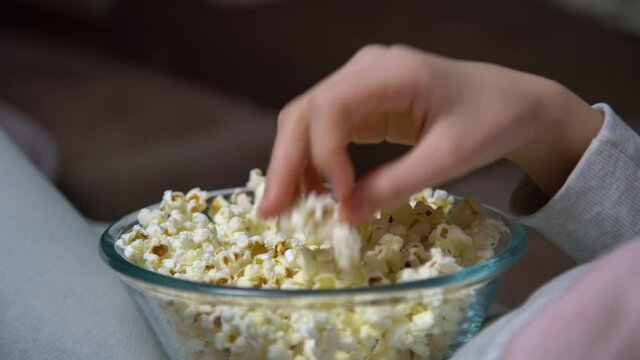 Close up of hand taking popcorn from a bowl while watching TV. Person sitting in comfortable couch and watching home cinema in the dark.