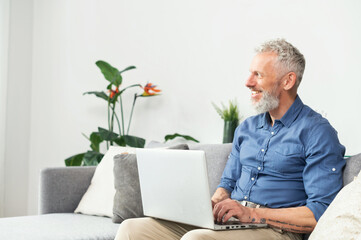 Smiling middle-aged hipster man in blue smart casual shirt using laptop sitting on the couch at home, working on new startup, senior gray haired journalist typing, texting, writing an article
