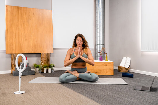 Full body of concentrated young female in activewear sitting in Lotus pose with closed eyes and prayer hands doing meditation during yoga session at home near smartphone placed on tripod