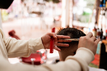 Crop anonymous male stylist with trimmer cutting hair of client in cape in barbershop