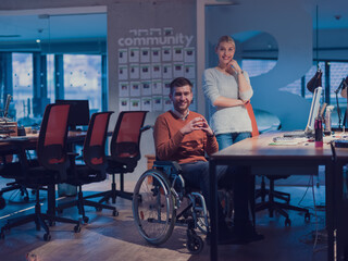 Fototapeta na wymiar Businessman in a wheelchair in modern coworking office space working late night in office. Colleagues in background. Disability and handicap concept. 