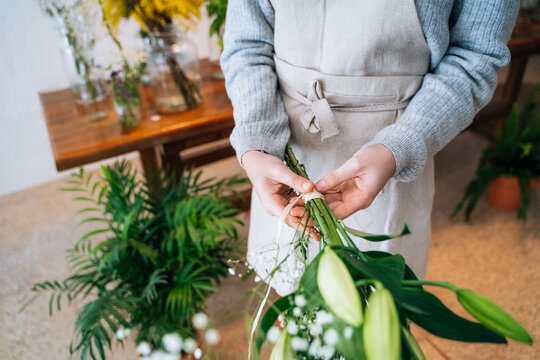 Cropped unrecognizable female florist in casual clothes and apron tying delicate bouquet of orange lily and white gypsophila in floral shop