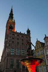 The Neptune's Fountain in Old Town of Gdansk, Poland at night. The fountain is lit with red and white lights. Town Hall building in the back. Dark night lit up with city lights. Night city tour.