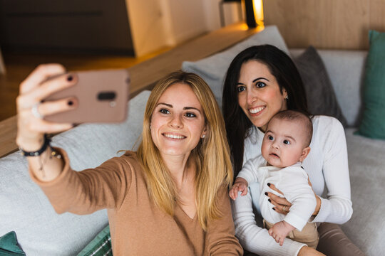 High angle of delighted LGBT family with adorable toddler sitting on couch and taking self shot on smartphone at home