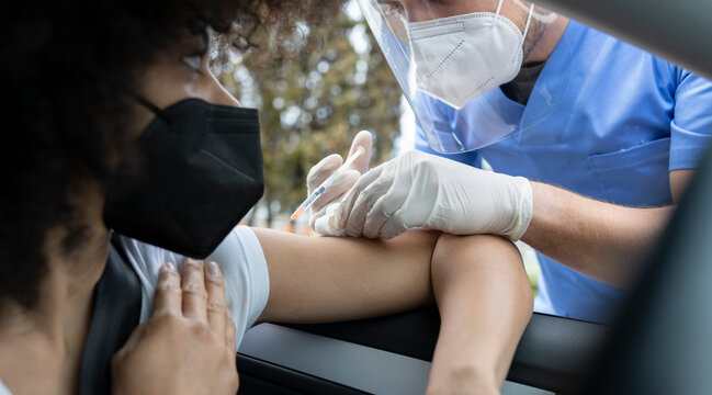 Side view of male doctor in protective uniform, latex gloves and face shield vaccinating African American female patient inside the car on a drive through mobile clinic during coronavirus outbreak