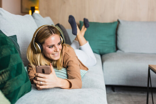 Joyful female lying on sofa and enjoying music in headphones while looking away using smartphone