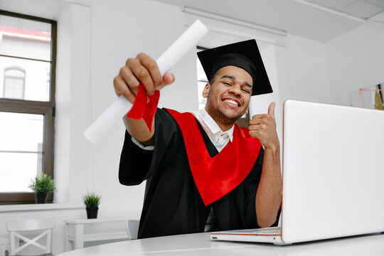 A Black student in graduate gown and square cap who is happy to finish his studies shows his the long-awaited diploma