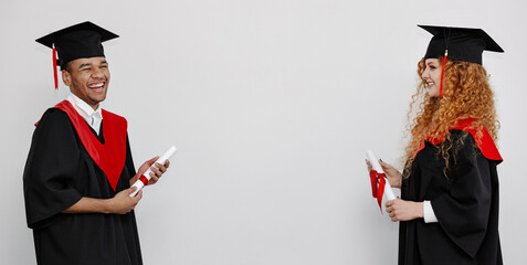 A black and redhaired students in graduation gowns and square caps stand opposite each other...