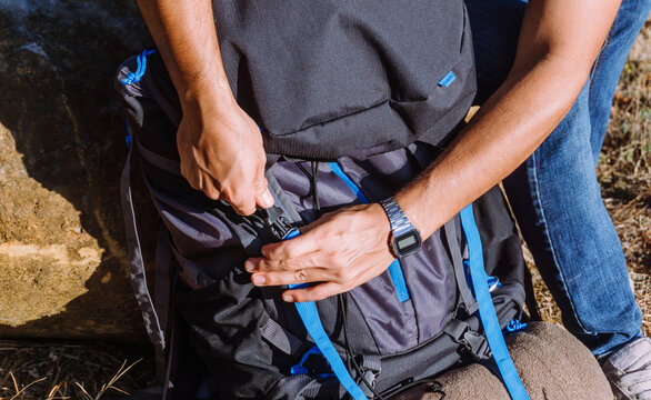 High angle of crop man in casual wear fastening backpack while trekking in sunny nature