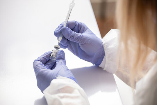 Unrecognizable medic in protective face shield mask and latex gloves with vial of coronavirus vaccine and syringe showing to camera while standing in hospital room