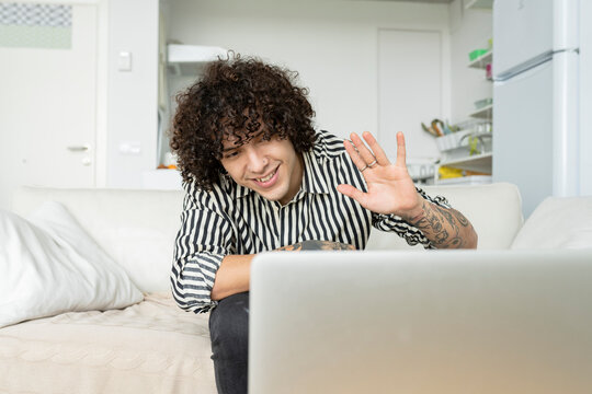 Content male with tattoos showing hi gesture against netbook during video chat while sitting on couch in apartment