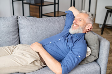 Serene grey-haired hipster guy rests on the couch in cozy living room, mature man lying down on the sofa, relaxing at home, smiling senior male reflecting, daydreaming on the sofa, has restful moment