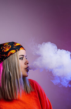 Side view of cool female in street style outfit smoking e cigarette and exhaling smoke through nose on purple background in studio with pink neon illumination