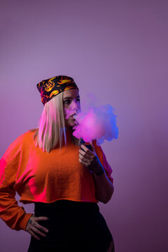 Cool female in street style outfit smoking e cigarette and exhaling smoke through nose and mouth on purple background in studio with pink neon illumination
