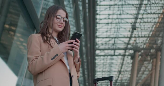 Charming business lady in eyeglasses with suitcase and smartphone standing near modern airport. Concept of people, business trip and technology. 