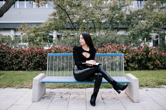 Stylish female entrepreneur sitting on bench and taking notes in organizer while working in urban park
