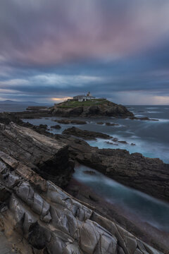 Breathtaking scenery of rocky island with lighthouse located in ocean near rocky coast in Faro Tapia de Casariego in Asturias in Spain under cloudy sky at sunrise