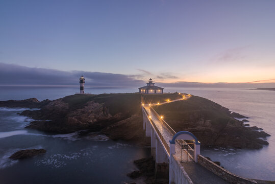 Spectacular scenery of bridge leading to rocky island covered with green grass with lighthouse placed in wavy ocean in Faro Illa Pancha in Galicia in Spain in twilight