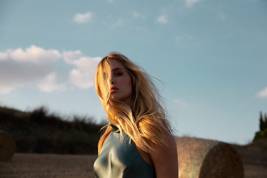 Peaceful female in elegant dress standing on dry field in rural area and looking at camera