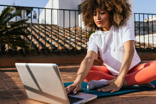 Focused African American female with curly hair choosing online tutorial on laptop while sitting on mat on rooftop and preparing for yoga lesson