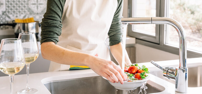 Crop young female in apron washing fresh strawberries under water fluid from tap in house kitchen