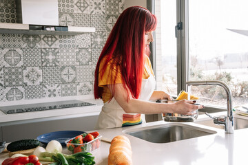 Young homosexual female pouring water from tap into saucepan with uncooked pasta against assorted vegetables in house
