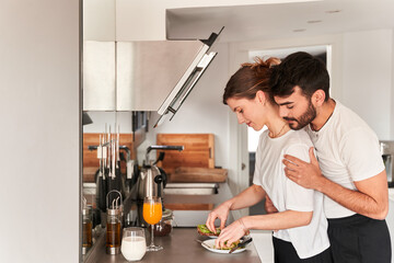 Side view of young romantic ethnic guy in white t shirt smiling and hugging happy girlfriend preparing healthy breakfast with avocado in kitchen