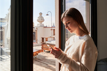 Side view of calm young lady in stylish sweater messaging on smartphone while standing near window at home