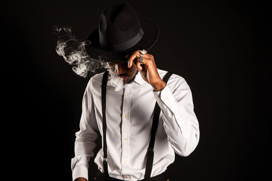 Masculine African American male in white shirt and hat exhaling vapor while smoking e cigarette