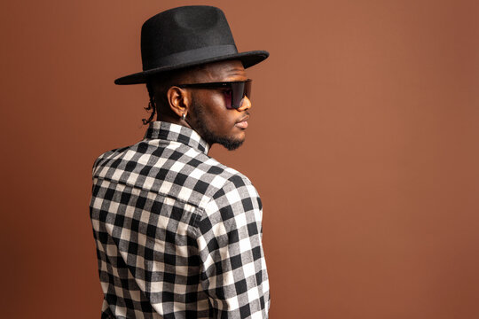 Young cool African American male model in checkered shirt, hat and sunglasses while standing on brown background