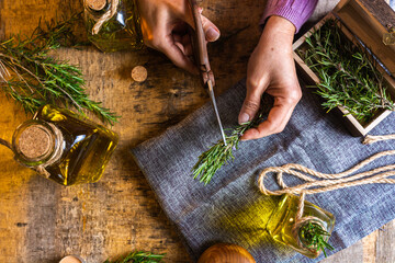 From above of crop lady cutting rosemary sprigs with scissors at table with fabric and rope near essential oil glass bottles and small chest