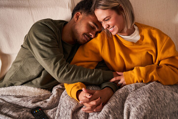 High angle of loving multiethnic couple relaxing on couch under blanket while cuddling and holding hands