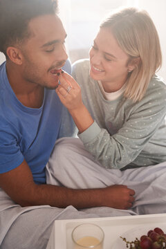 High angle of multiethnic couple in love feeding each other while sitting on bed and having tasty breakfast in morning