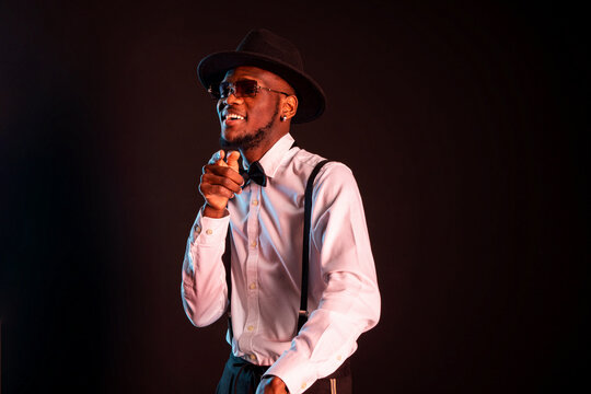 Young masculine ethnic male model in hat and trousers standing dancing while looking at camera on black background