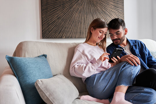 Romantic young diverse couple in casual clothes relaxing on sofa and watching photos on smartphone during weekend at home