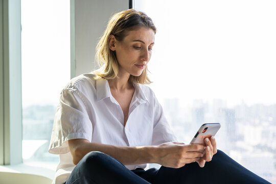 Lonely unemotional young woman sitting in empty office with big window browsing on the mobile phone