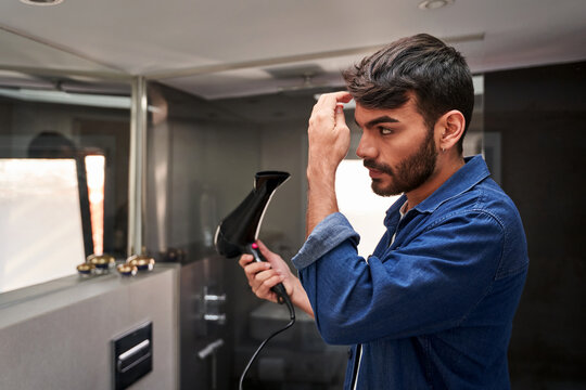 Side view of self assured young bearded Hispanic guy in denim shirt using hairdryer while adjusting hair in bathroom