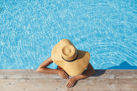 Enjoying summer vacation. Stylish woman in hat relaxing in pool water at wooden pier, top view. Slim young female sunbathing at  swimming pool edge. Travel and Holidays. Space for text
