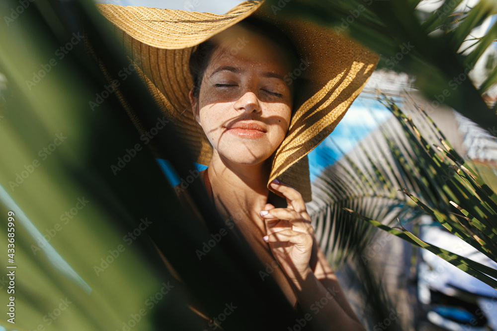 Wall mural portrait of beautiful carefree woman in hat relaxing under palm leaves at tropical resort, enjoying 