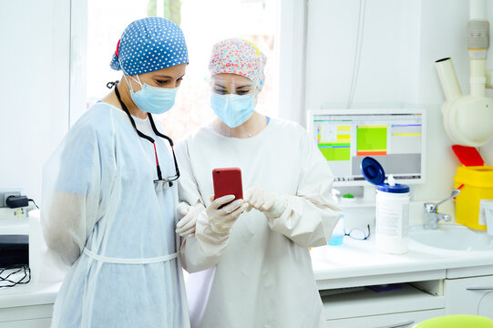 Female surgeons in medical cloth caps surfing internet on cellphone against desktop computer in light hospital